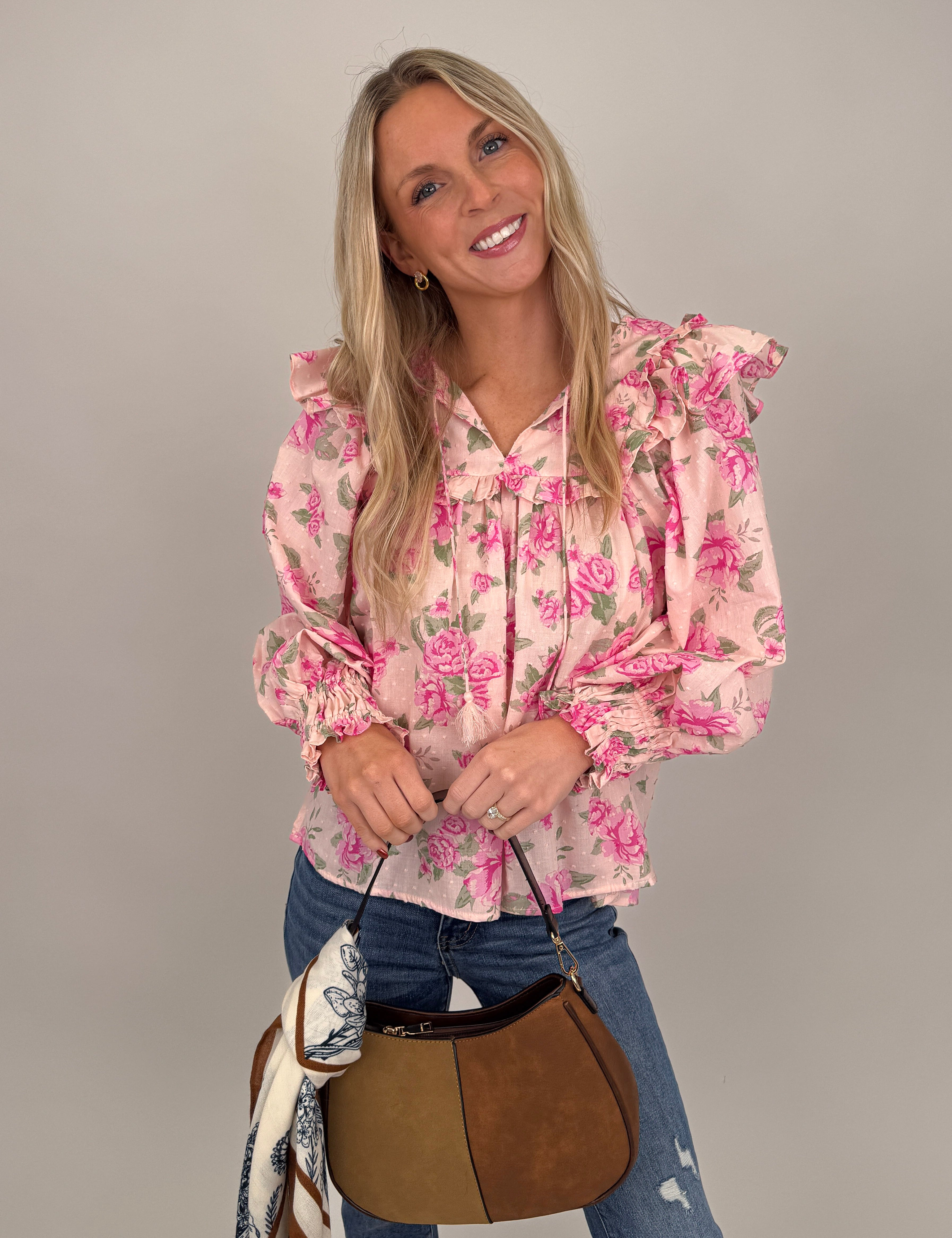 Woman wearing pink Amber Leigh Top with bold rose floral print, statement ruffle shoulders, swiss dot texture, and tassel tie neckline