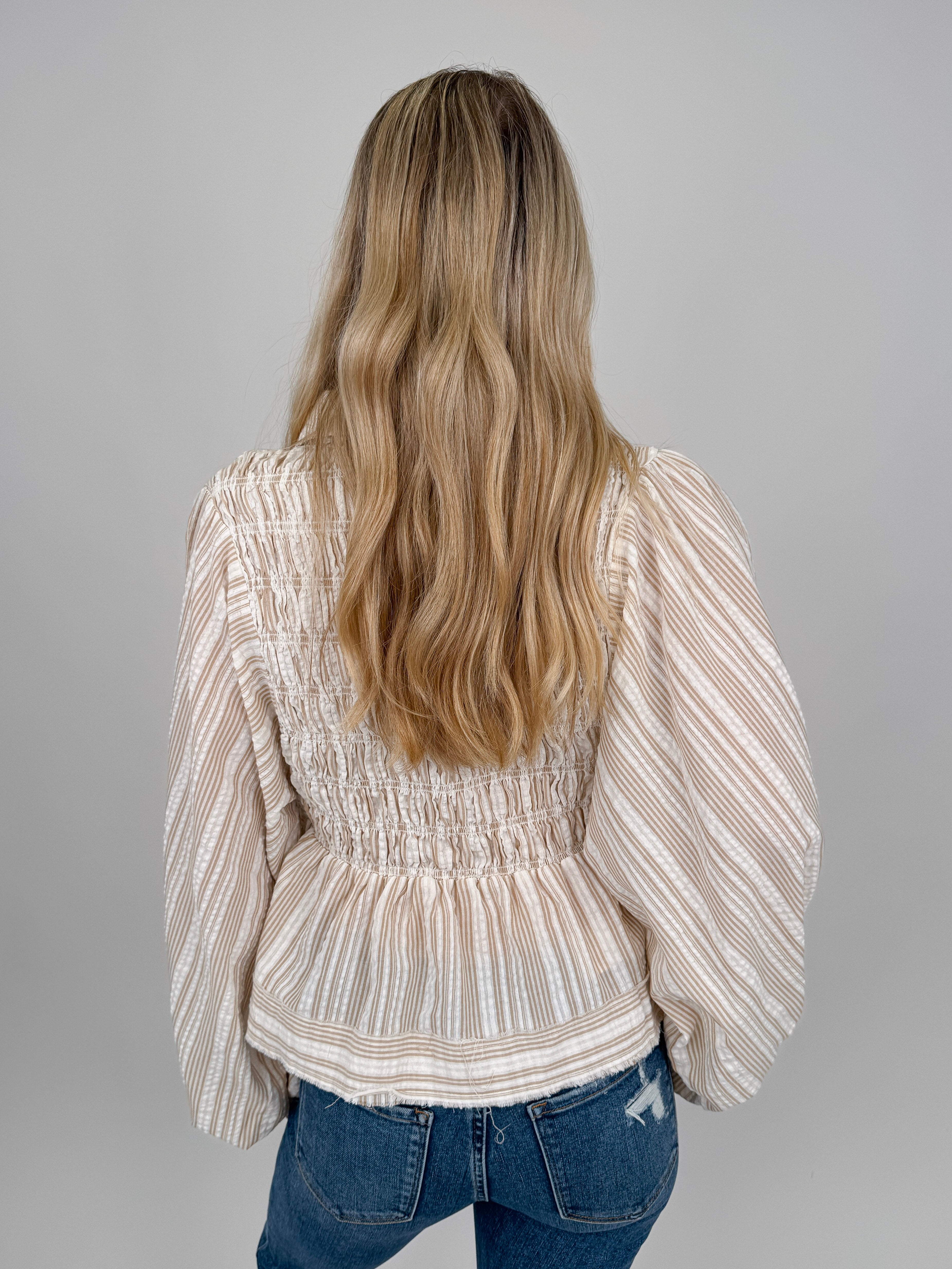 Woman wearing neutral striped Boardwalk Blouse with smocked bodice, collared V-neckline, billowy long sleeves, and relaxed flowing fit