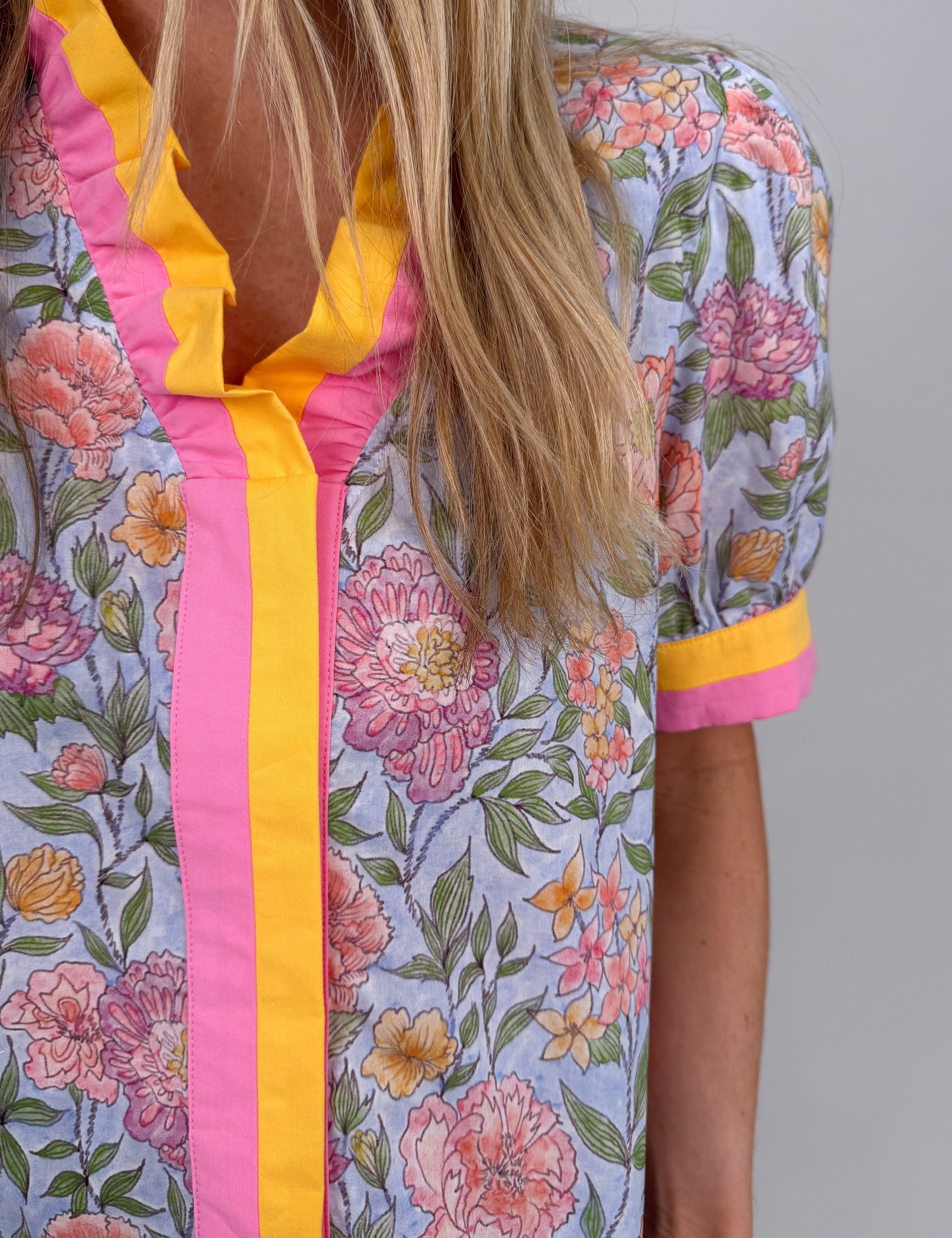 Woman wearing blue Sherbet Fields Floral Blouse with coral, pink, and yellow flowers, yellow-pink striped trim, puff sleeves, and ruffle V-neckline