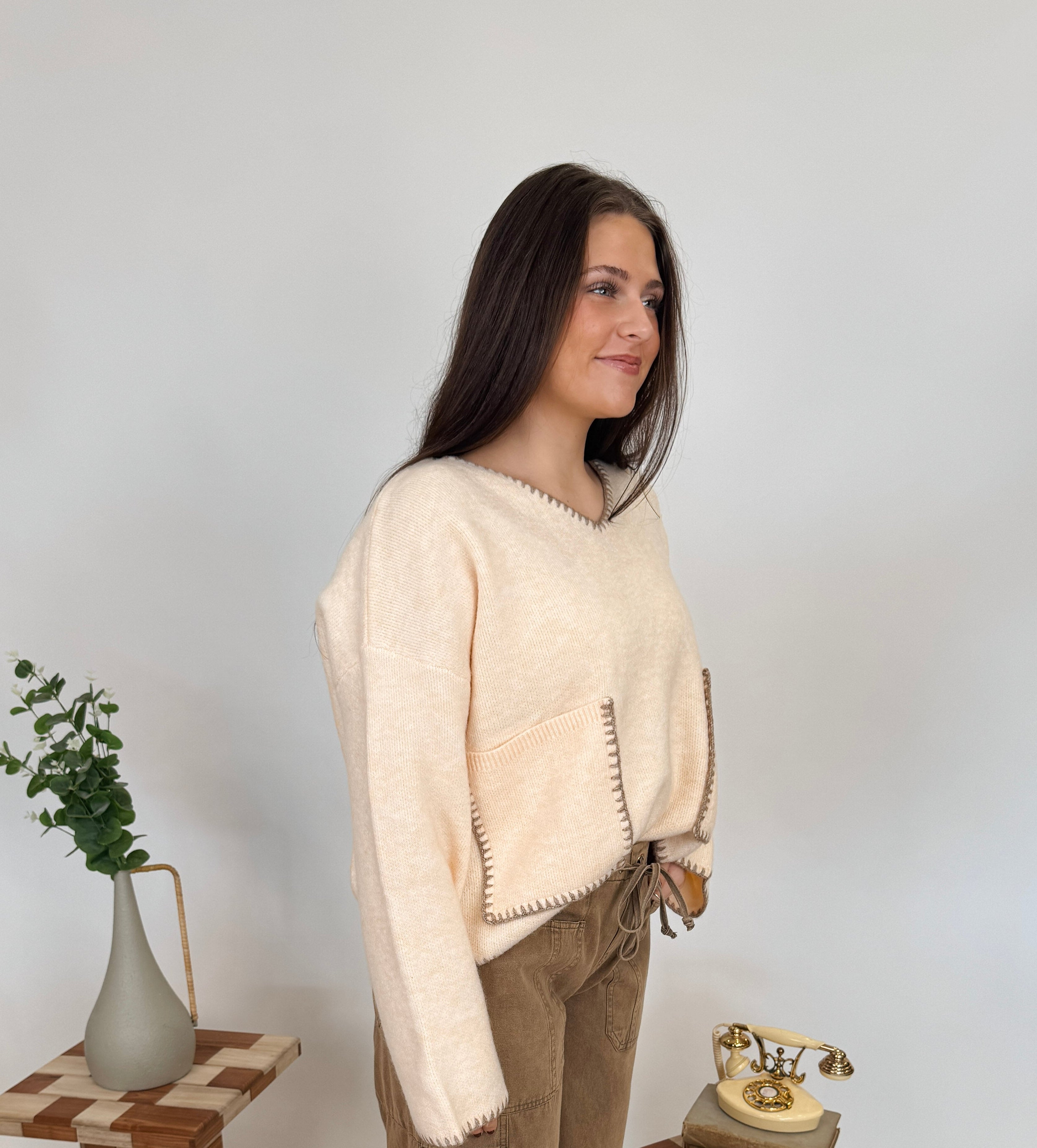 Woman in a beige sweater and brown pants standing in a room with decorative elements.