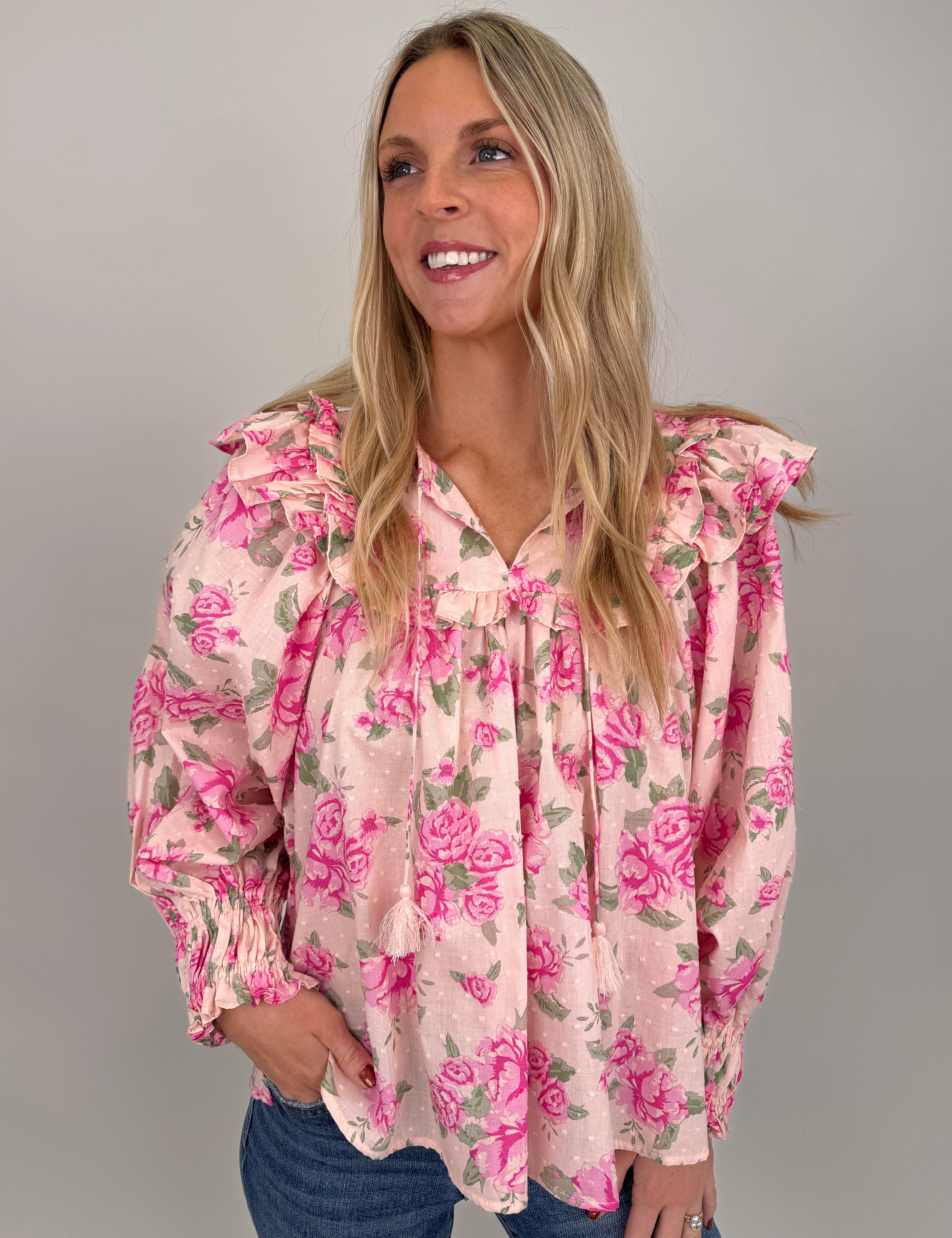 Woman wearing pink Amber Leigh Top with bold rose floral print, statement ruffle shoulders, swiss dot texture, and tassel tie neckline