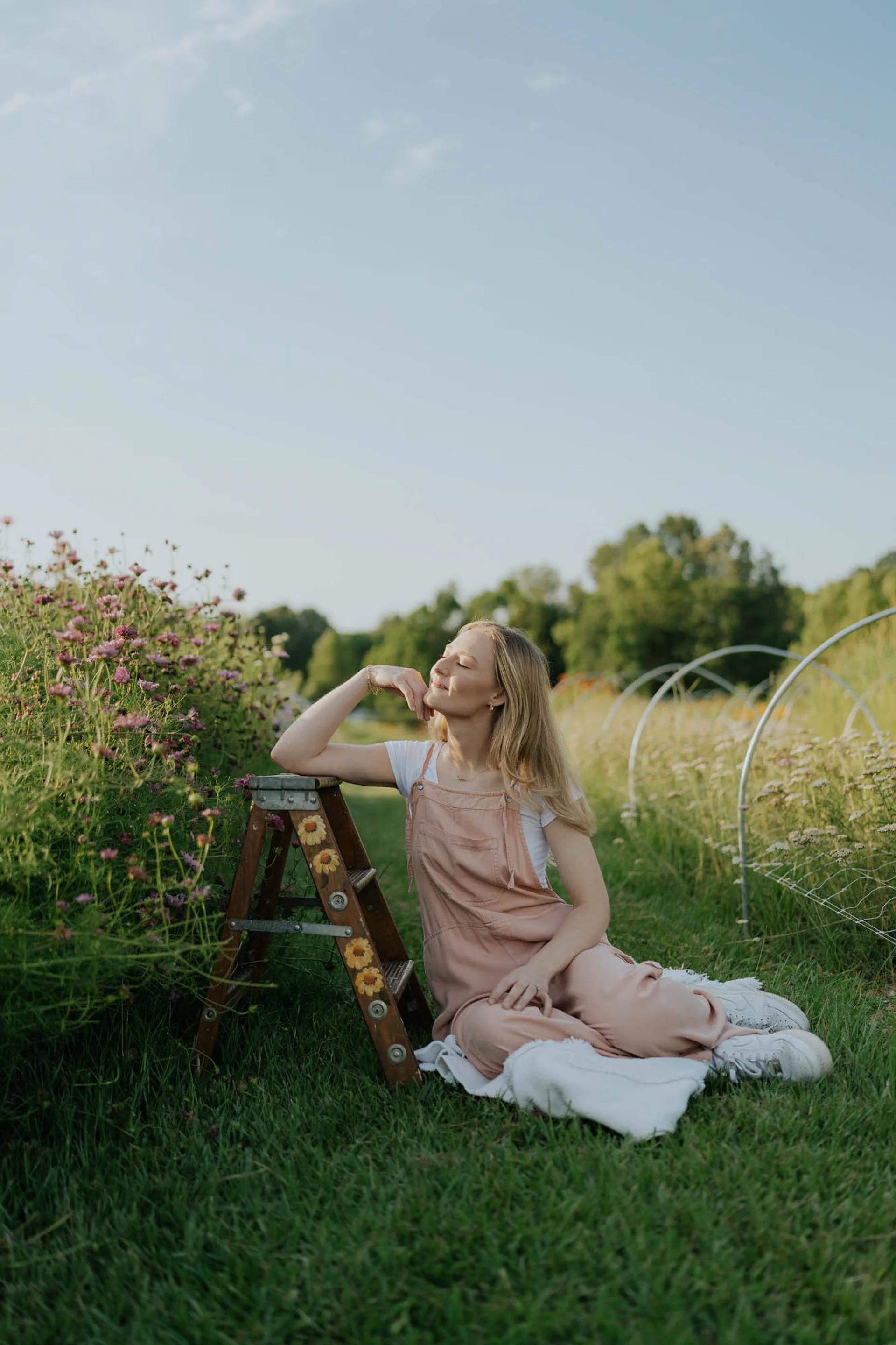 Woman in a pink dress sitting on a blanket in a field with a ladder and flowers nearby.