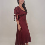 Katrina Midi Dress in burgundy crinkled satin with lace trim at the sweetheart neckline and empire waist, puff sleeves with bow ties, and a flowing bias cut midi skirt