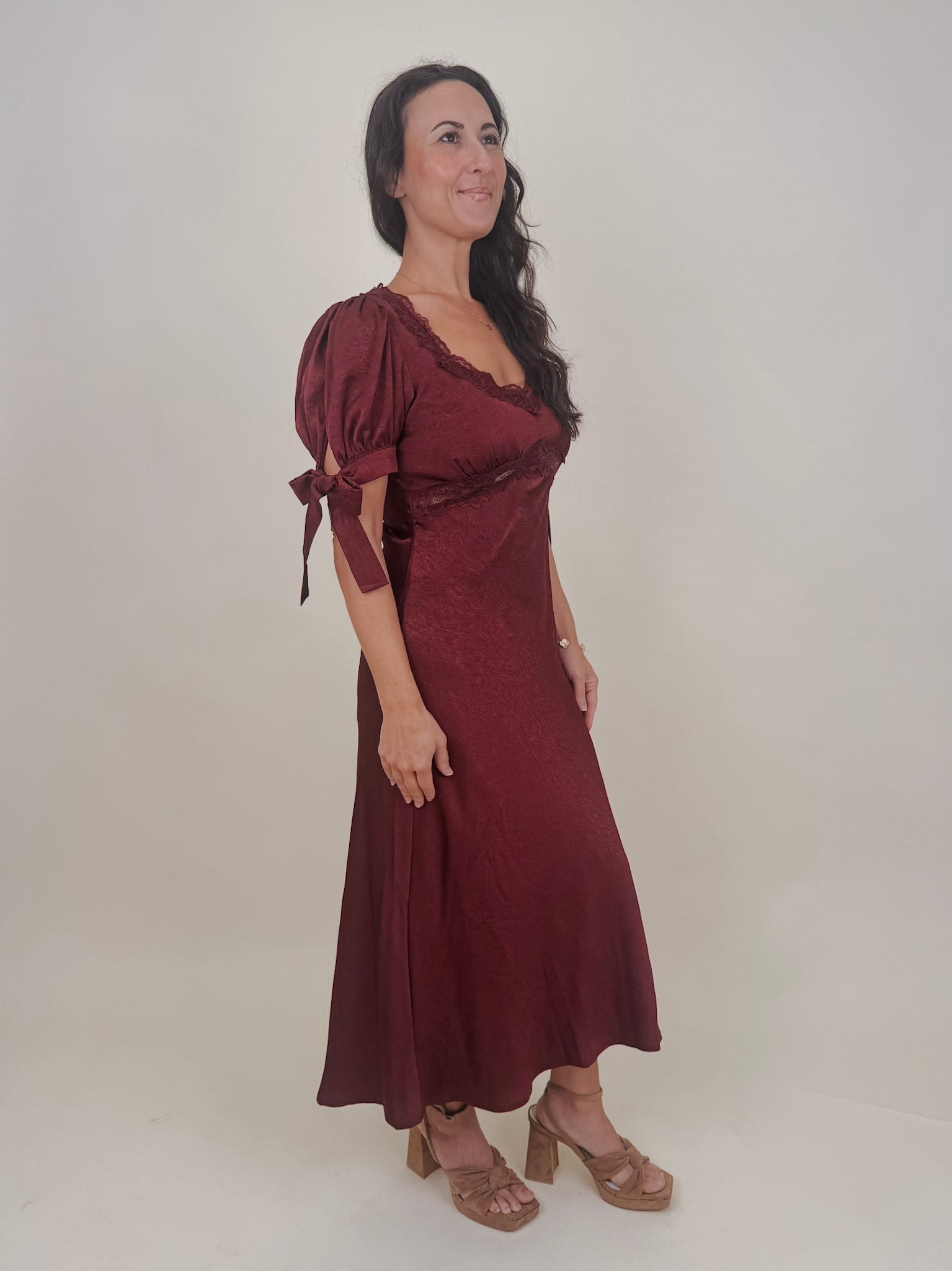 Katrina Midi Dress in burgundy crinkled satin with lace trim at the sweetheart neckline and empire waist, puff sleeves with bow ties, and a flowing bias cut midi skirt