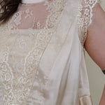 So Long London Blouse in champagne satin with eyelash lace panels, a mock neckline, cap sleeves, and a ruffled peplum hem with a ribbon tie back