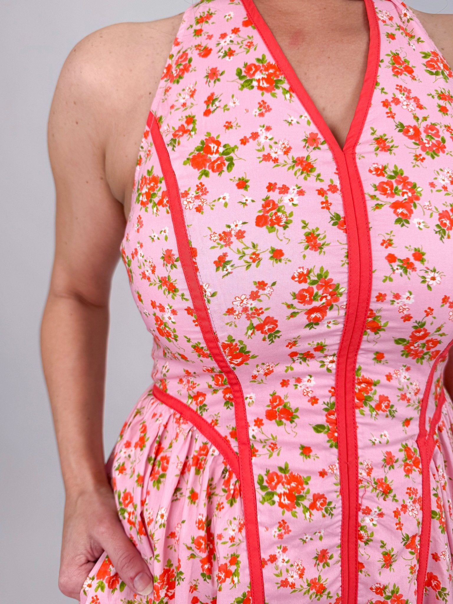 Tara Dress in pink ditsy floral print with red contrast piping, halter V neckline, fitted bodice, and full gathered midi skirt