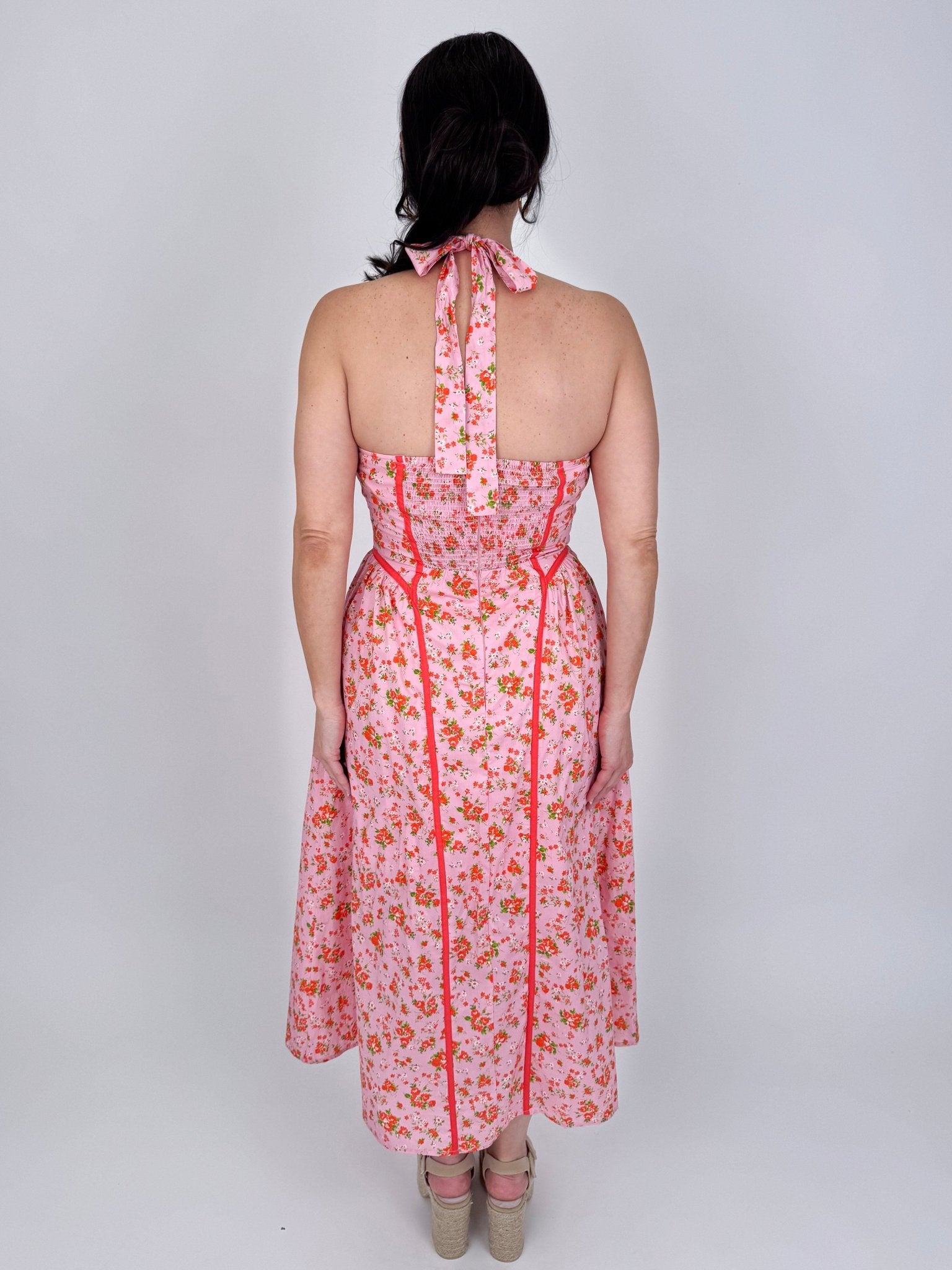 Tara Dress in pink ditsy floral print with red contrast piping, halter V neckline, fitted bodice, and full gathered midi skirt